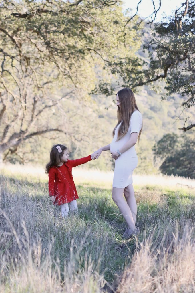 Pregnant mom holding her daughter's hand as they stand in a field and smile at each other