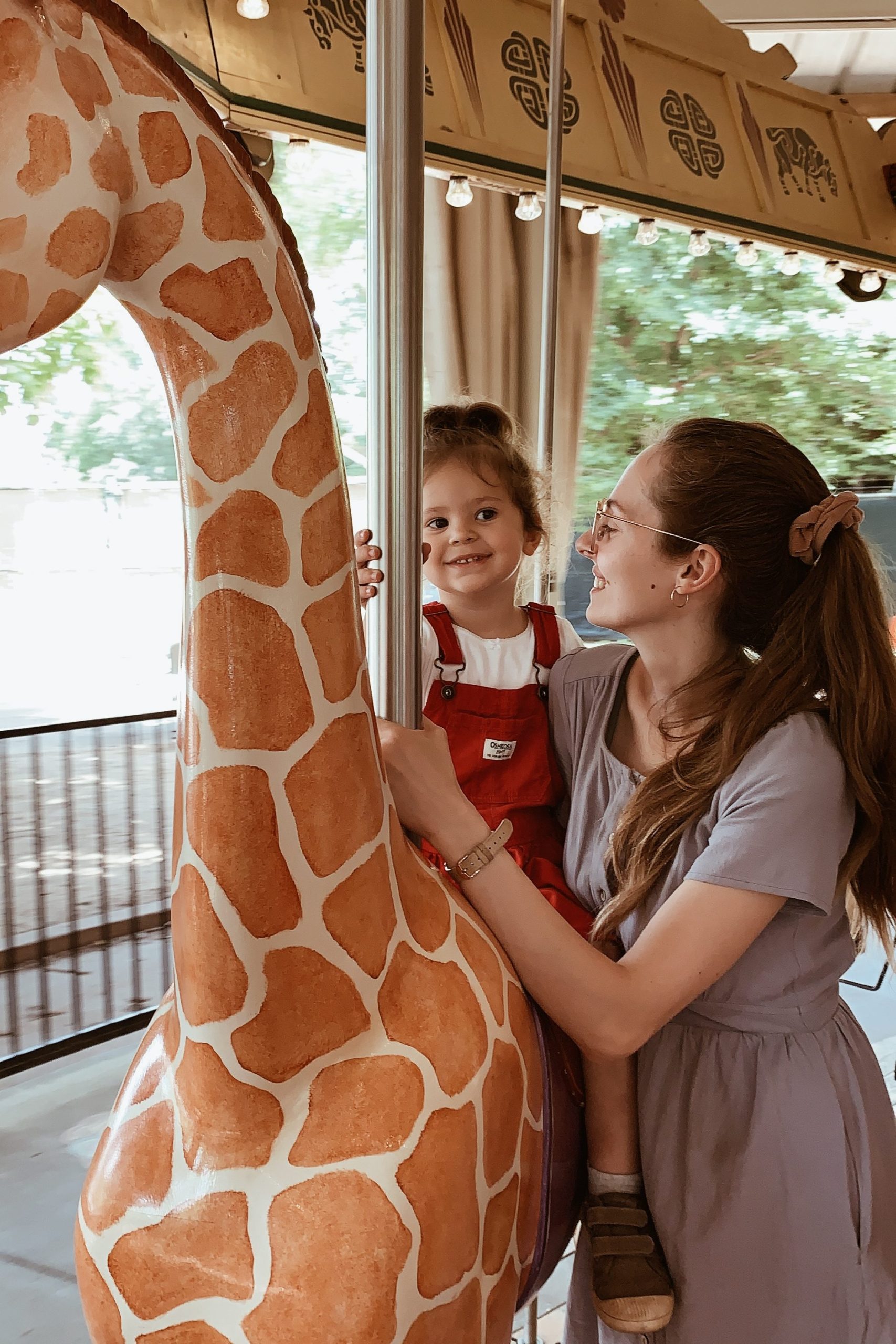 Mother is holding her toddler as her toddler sits on a giraffe on a carousel.