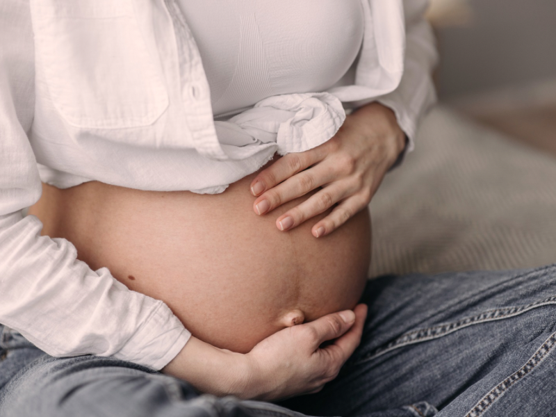 A pregnant woman is sitting on her bed, embracing her belly, wearing a white shirt tied in a knot above her belly, and jeans under her belly