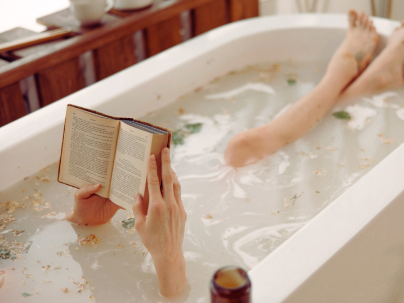 A woman is taking a bath and reading a book. The bath has flowers floating.